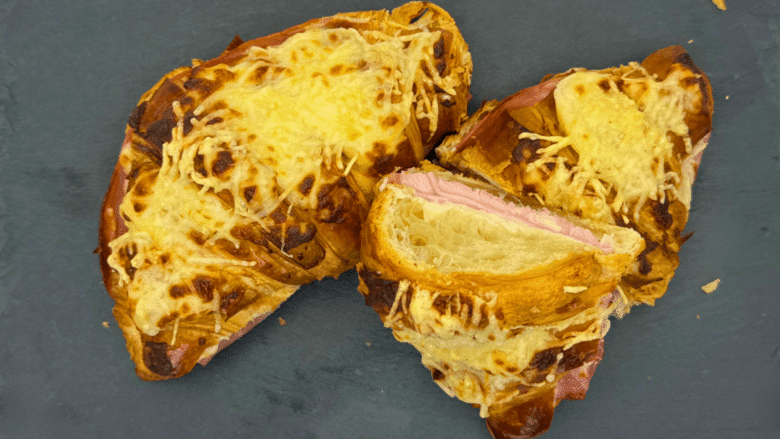 Croissants au jambon et fromage gratinés. Deux croissants dorés, garnis de jambon et de fromage fondant, présentés sur une ardoise noire. Parfait pour un dîner rapide et savoureux. Recette facile à réaliser à la maison.